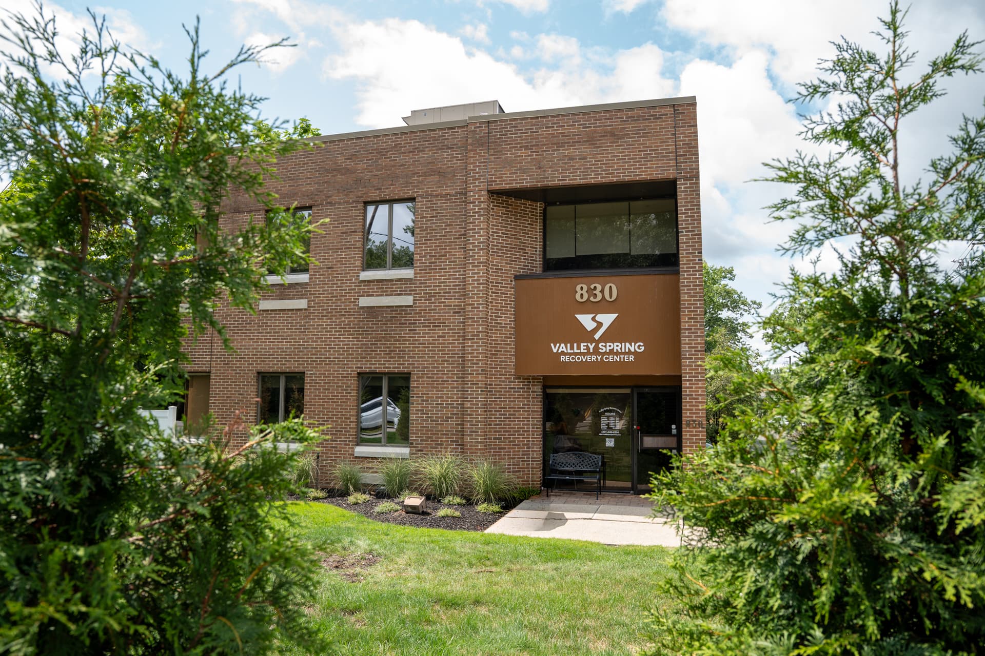 The Valley Spring Recovery Center entrance at 830 Broadway, Norwood, New Jersey — the brick building with the 830 and Valley Spring signage, framed by evergreens with a bench beside the front door