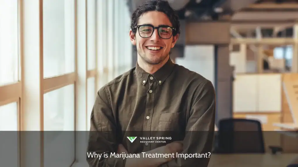 Young man with glasses smiles confidently while standing near large windows in a modern office setting, embodying a welcoming atmosphere.