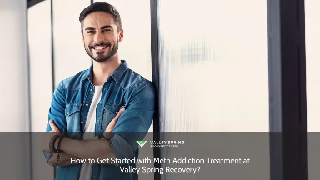 Smiling man with crossed arms, wearing a denim shirt, leans against a glass wall, embodying confidence. Ideal for discussing recovery at Valley Spring Recovery.